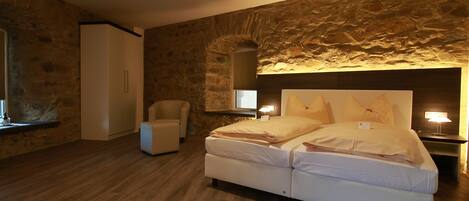 Un dormitorio con pared de piedra, una cama, un sillón y una mesita de noche con lámpara.