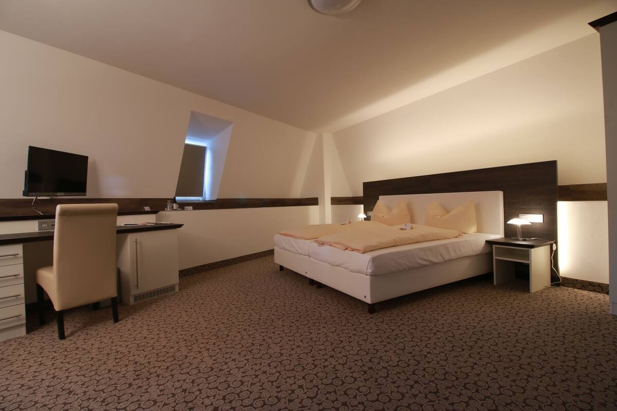 A hotel room with a bed, desk, chair, TV, and a small table with a lamp.
