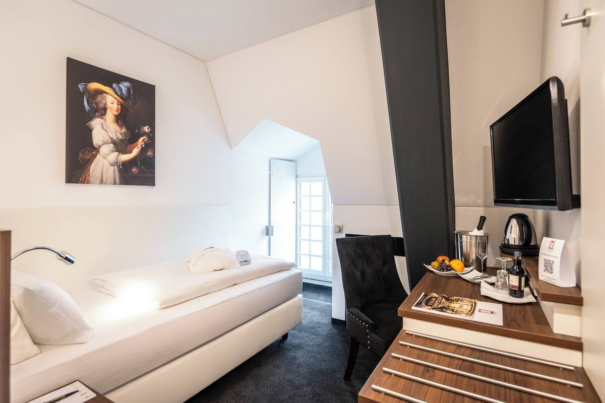 A modern hotel room with a bed, a desk with a coffee maker, a TV, and a painting on the wall.
