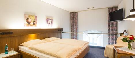 A hotel room with a bed, bedside table, a desk with a chair, a TV mounted on the wall, and a window with curtains.