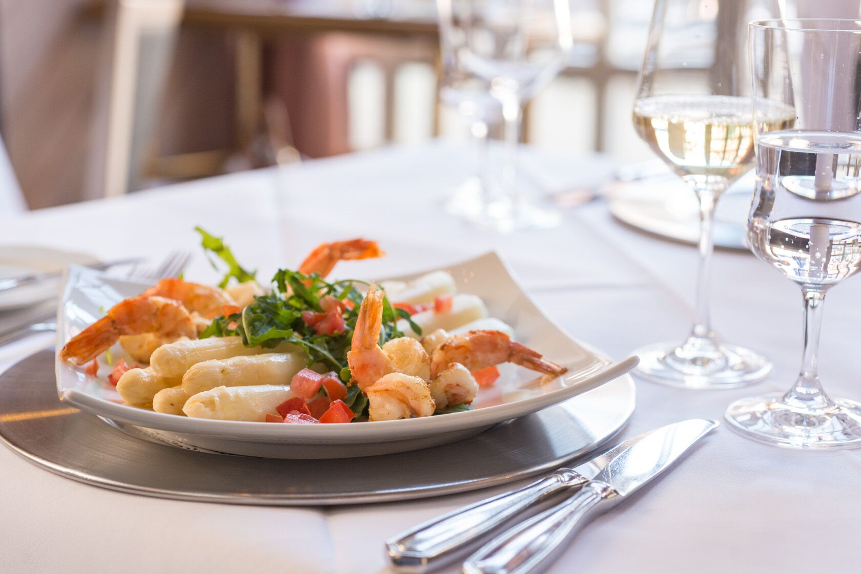 Un plato de comida con camarones y vegetales, acompañado de un tenedor y cuchillo sobre una mesa con una copa de vino blanco.