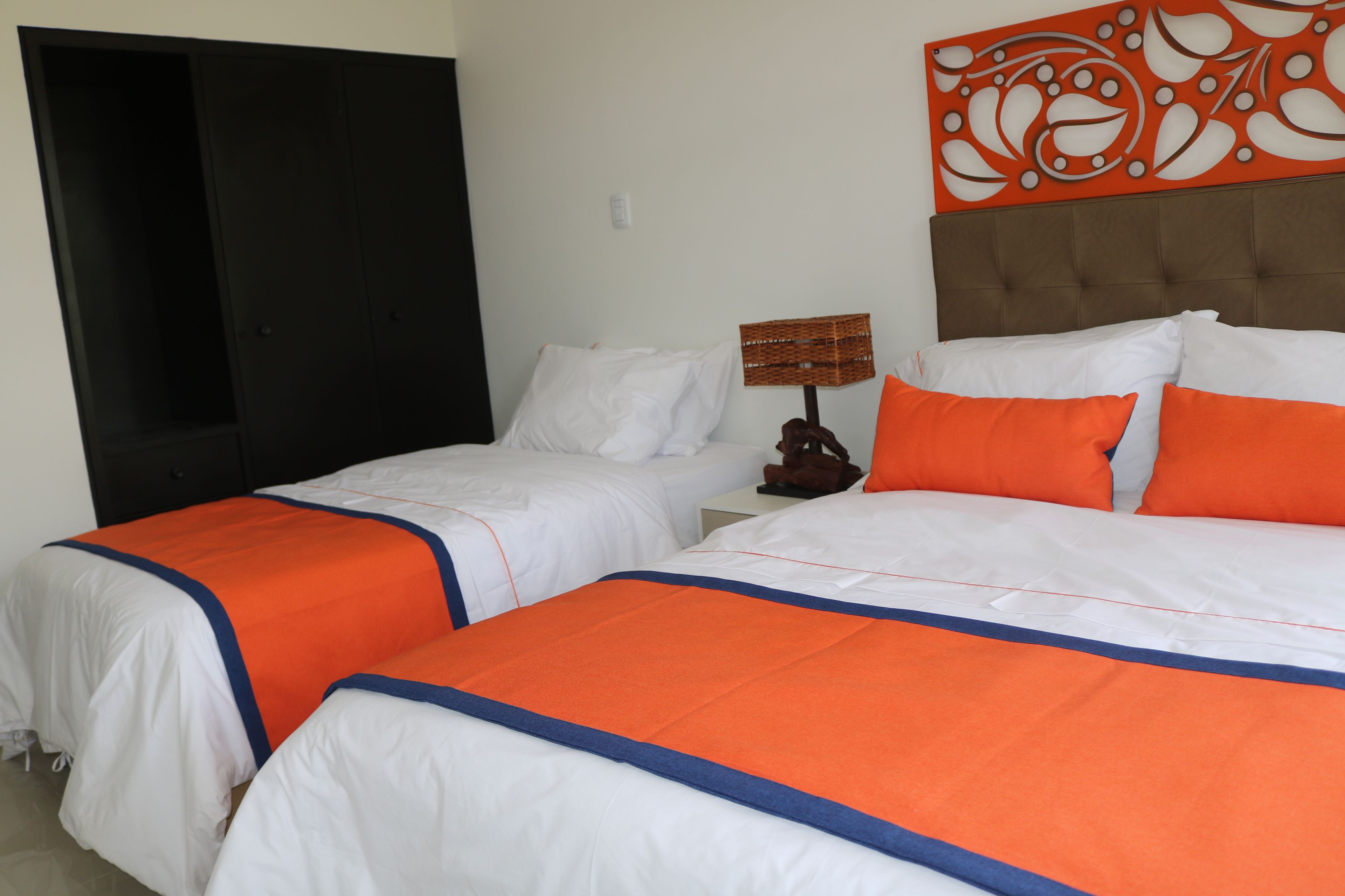 Habitación de hotel con dos camas, cada una con ropa de cama blanca y detalles anaranjados, cabeceros marrones y un adorno de pared decorativo.