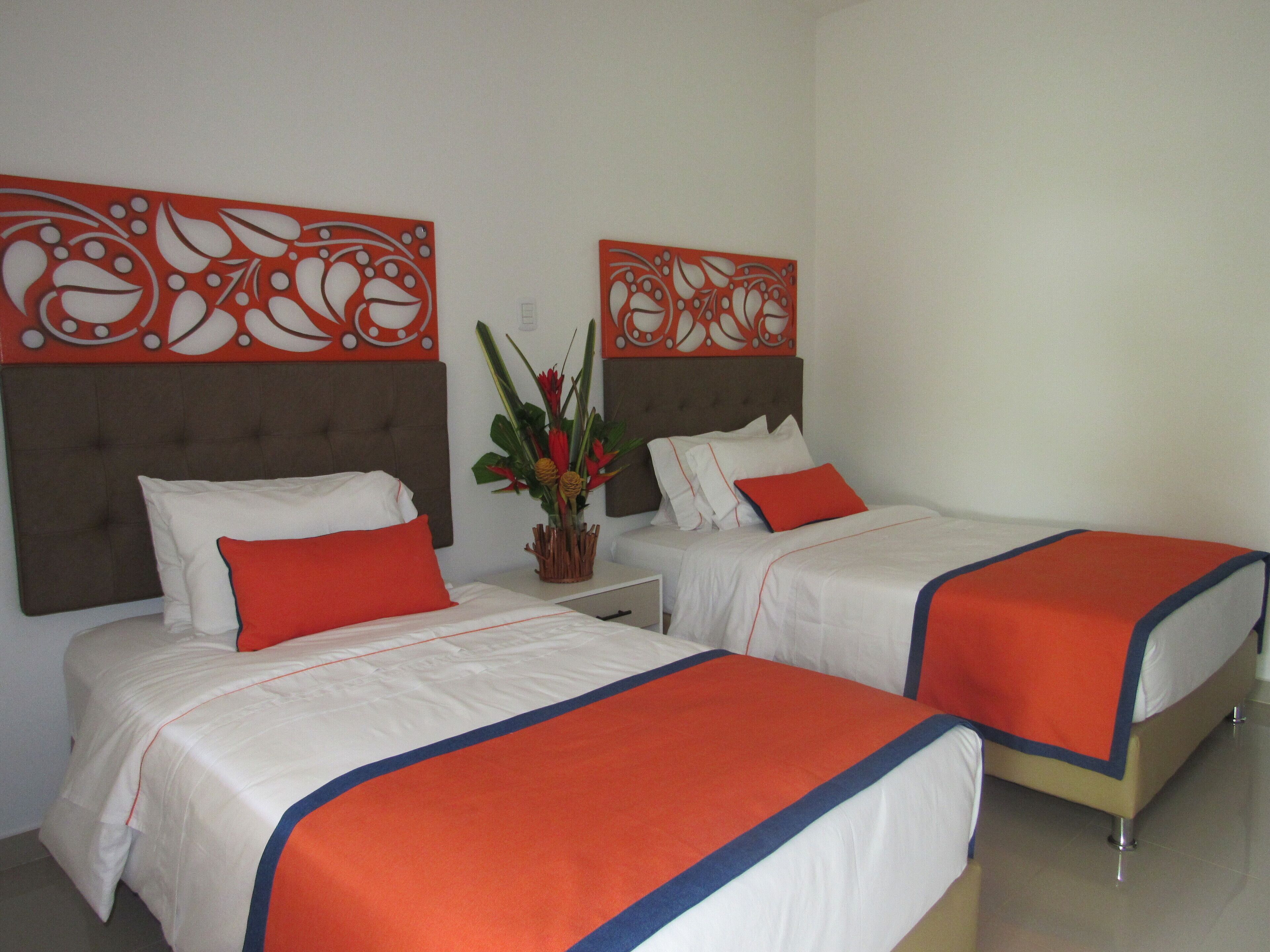 A hotel room with two beds, each with white bedding and orange accents, a decorative wall panel, and a small plant on the nightstand.