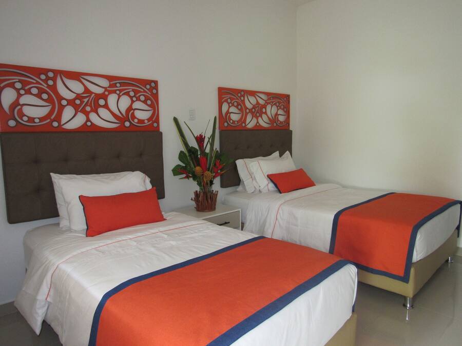 A hotel room with two beds, each with white bedding and orange accents, a decorative wall panel, and a small plant on the nightstand.