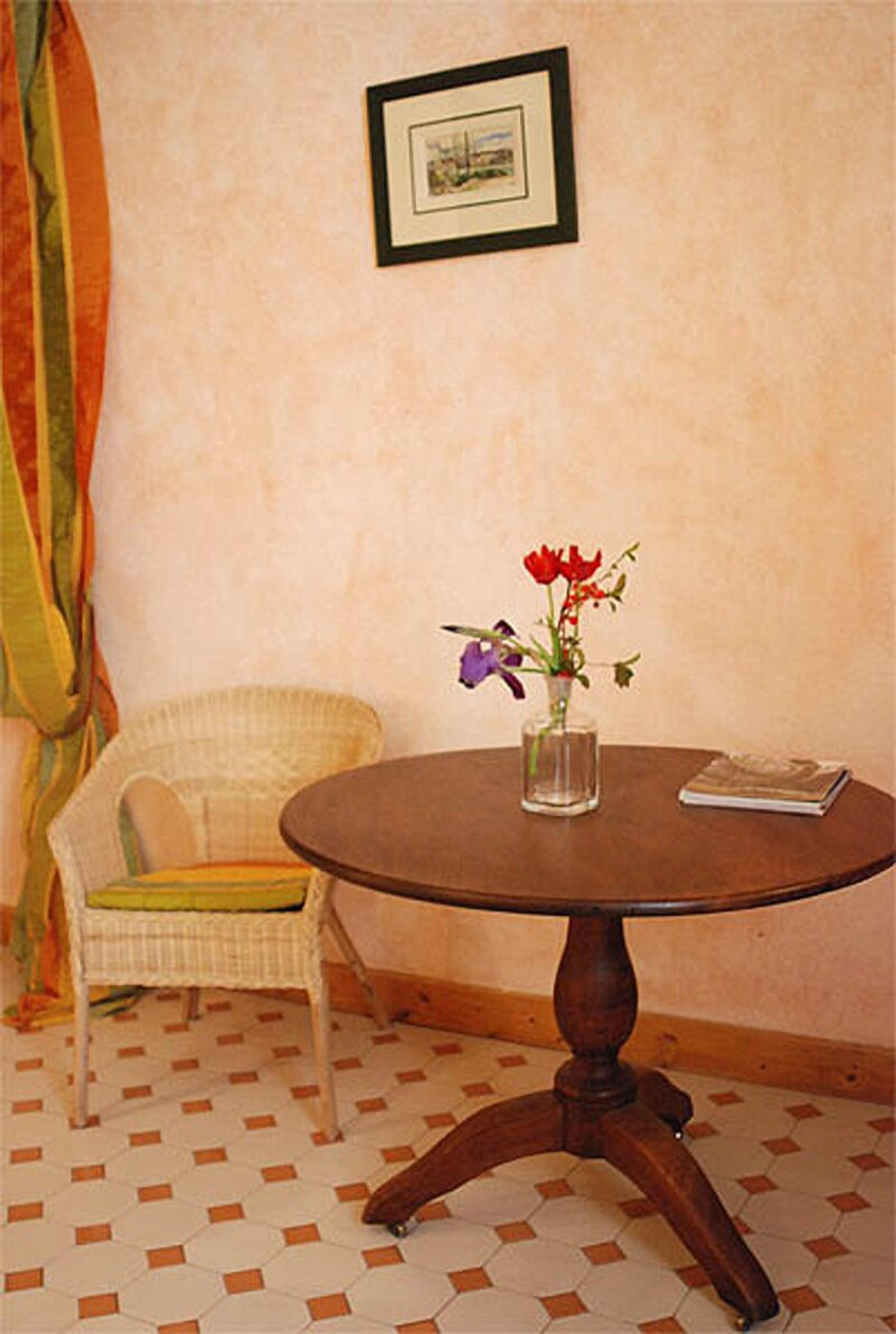 Une table ronde en bois, sur laquelle est posé un vase de fleurs, une chaise avec un coussin et un tableau encadré accroché au mur.