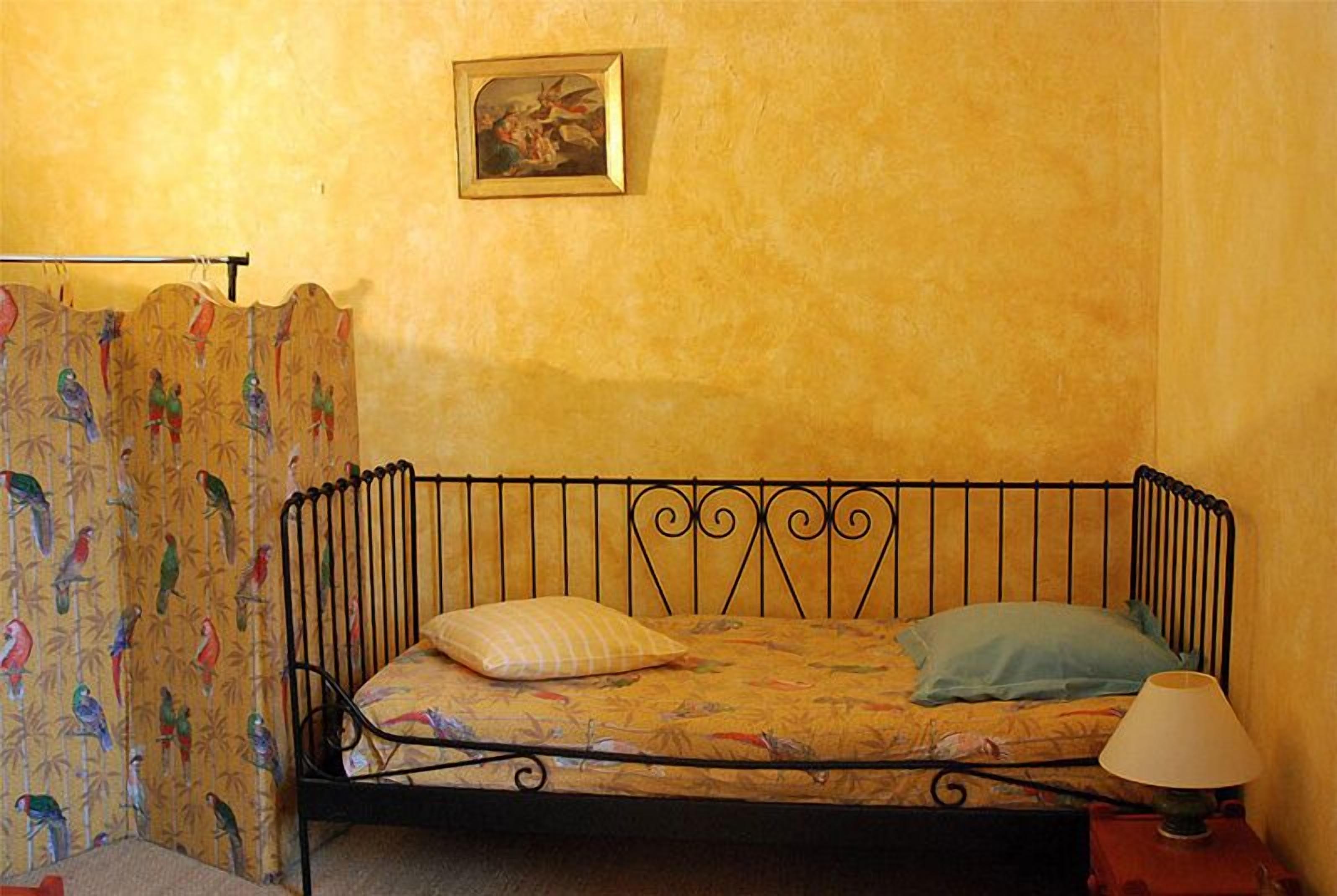 Een enkel bed met een metalen frame, een beddegoed met een patroon en twee kussens. Naast het bed staat een klein tafeltje met een lamp.
