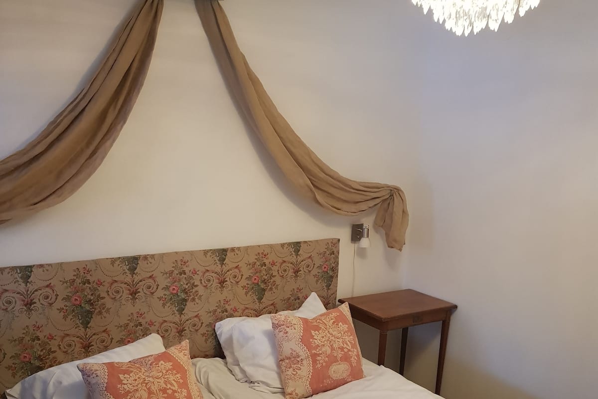 A double bed with floral headboard, two pillows, a nightstand, and a chandelier.
