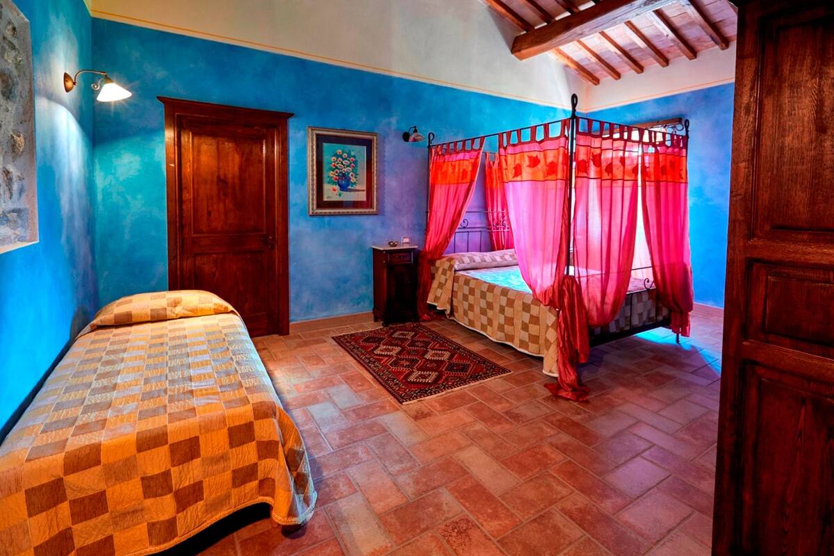 A bedroom with a checkered bedspread, a wooden door, a framed painting, a red canopy bed, and a patterned rug.