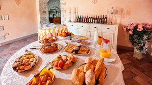 Ein Frühstücksbuffet mit verschiedenen Backwaren, Früchten und Brot auf einem Tisch in einem Raum mit Ziegelwand und einem weißen Schrank mit Weinflaschen.