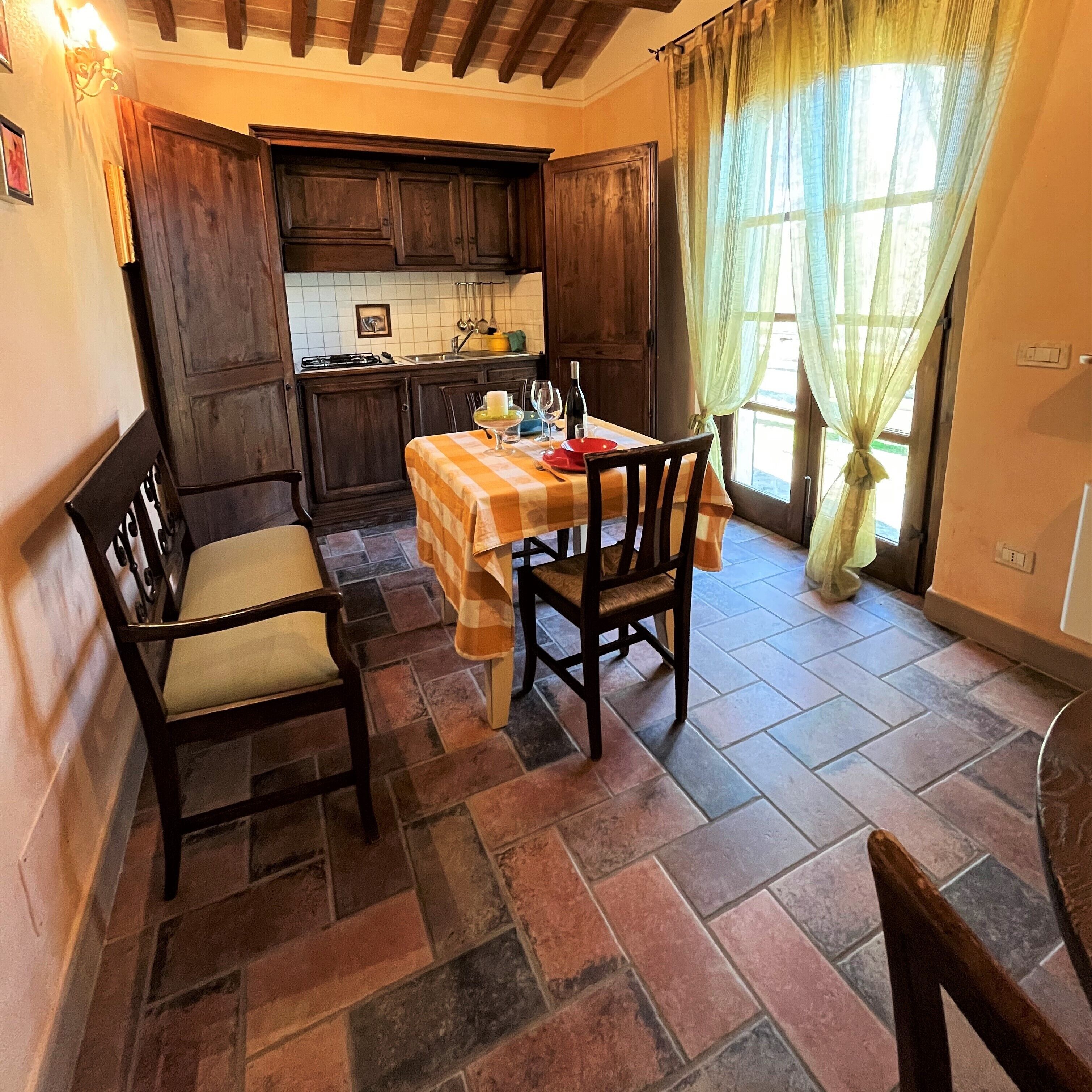 A kitchen with a dining table set for two, wooden chairs, a tiled floor, and a window with curtains.