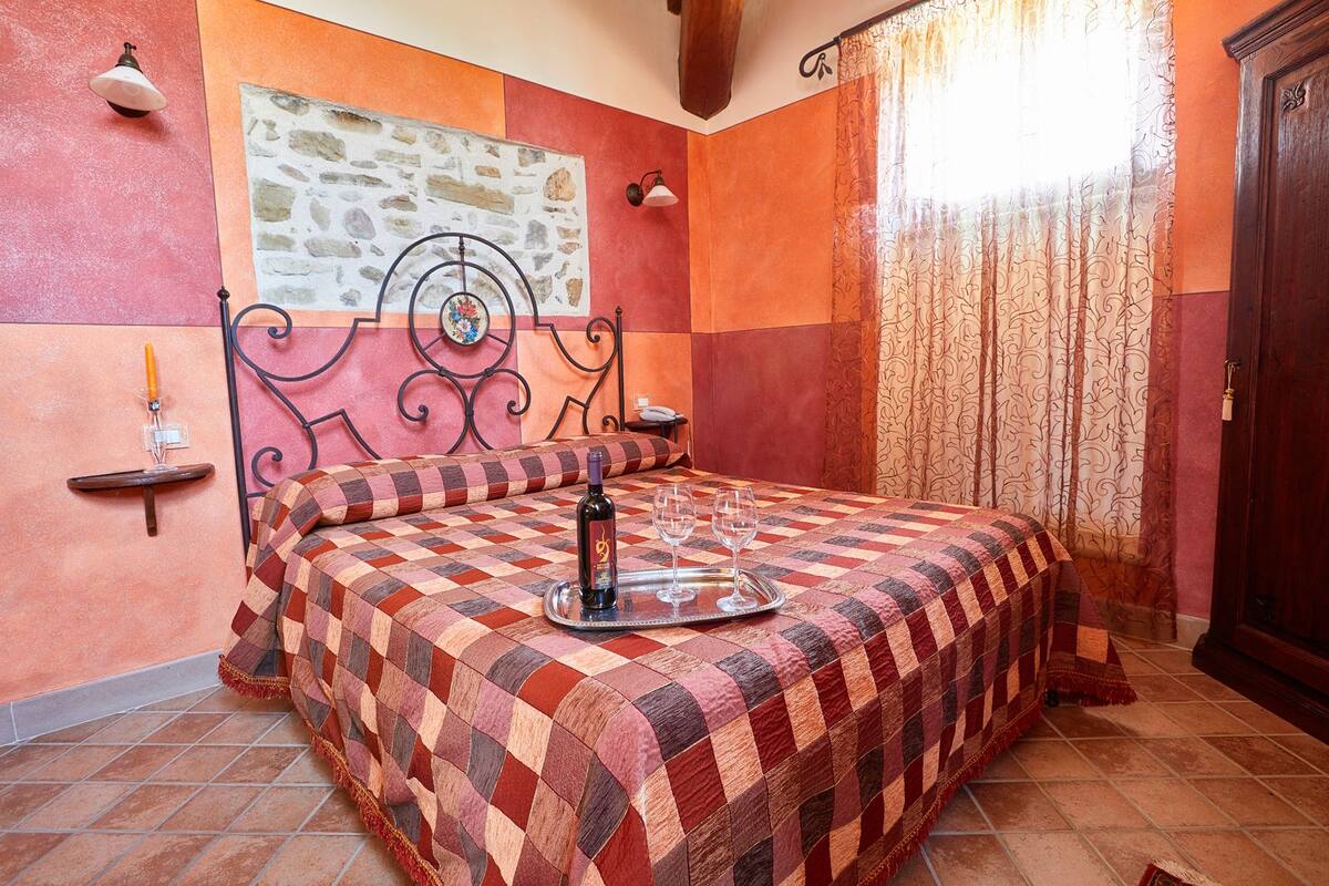 A bedroom with a checkered bedspread, a stone wall, a decorative metal headboard, and a window with patterned curtains.