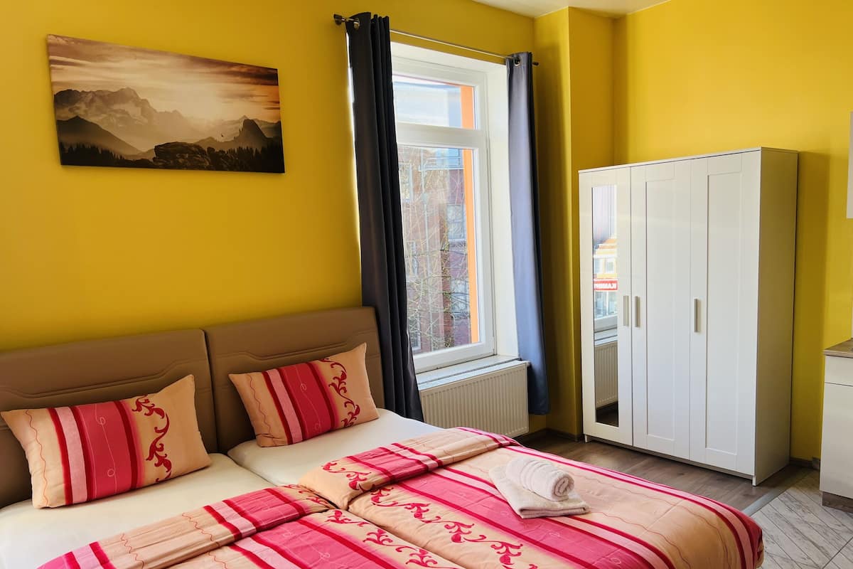 A bedroom with a bed, pillows, a window with curtains, a radiator, and a wardrobe.