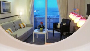 Habitación de hotel con sofá, una mesita con una planta, una silla y vista al mar a través de amplias ventanas.