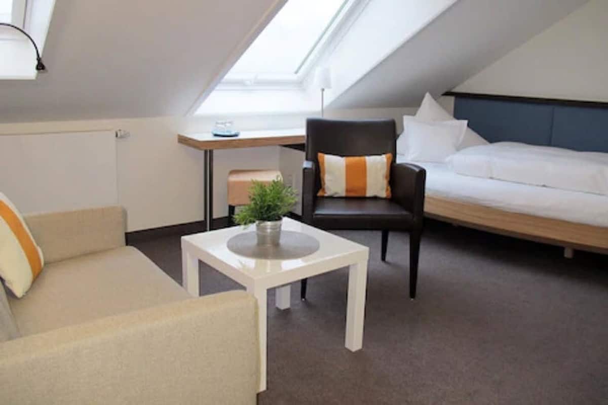 Quarto moderno e compacto com cama estilo loft, um sofá, uma pequena mesa com uma planta e uma escrivaninha com abajur.