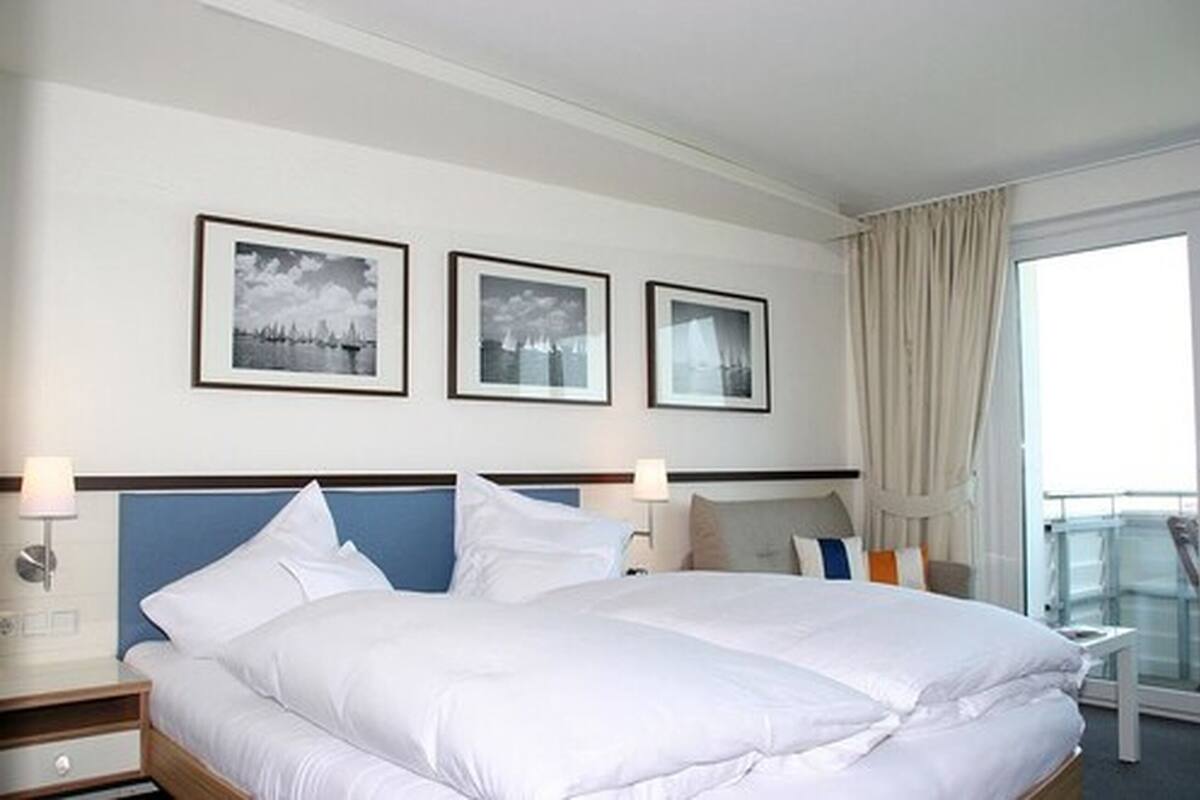Quarto de hotel com uma cama grande, duas mesas de cabeceira, uma janela com cortinas e três quadros na parede.