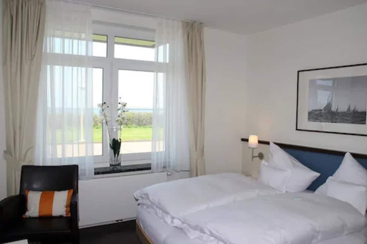Quarto de hotel com cama, poltrona, janela com cortinas, luminária na parede e quadro decorativo.