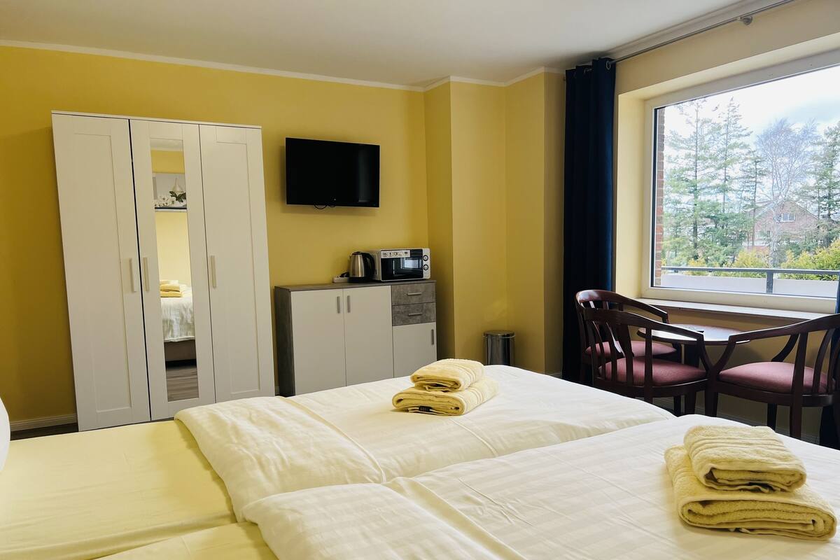 Une chambre d’hôtel équipée d’un lit, d’un bureau, d’une chaise, d’une télévision et d’une fenêtre donnant sur des arbres.