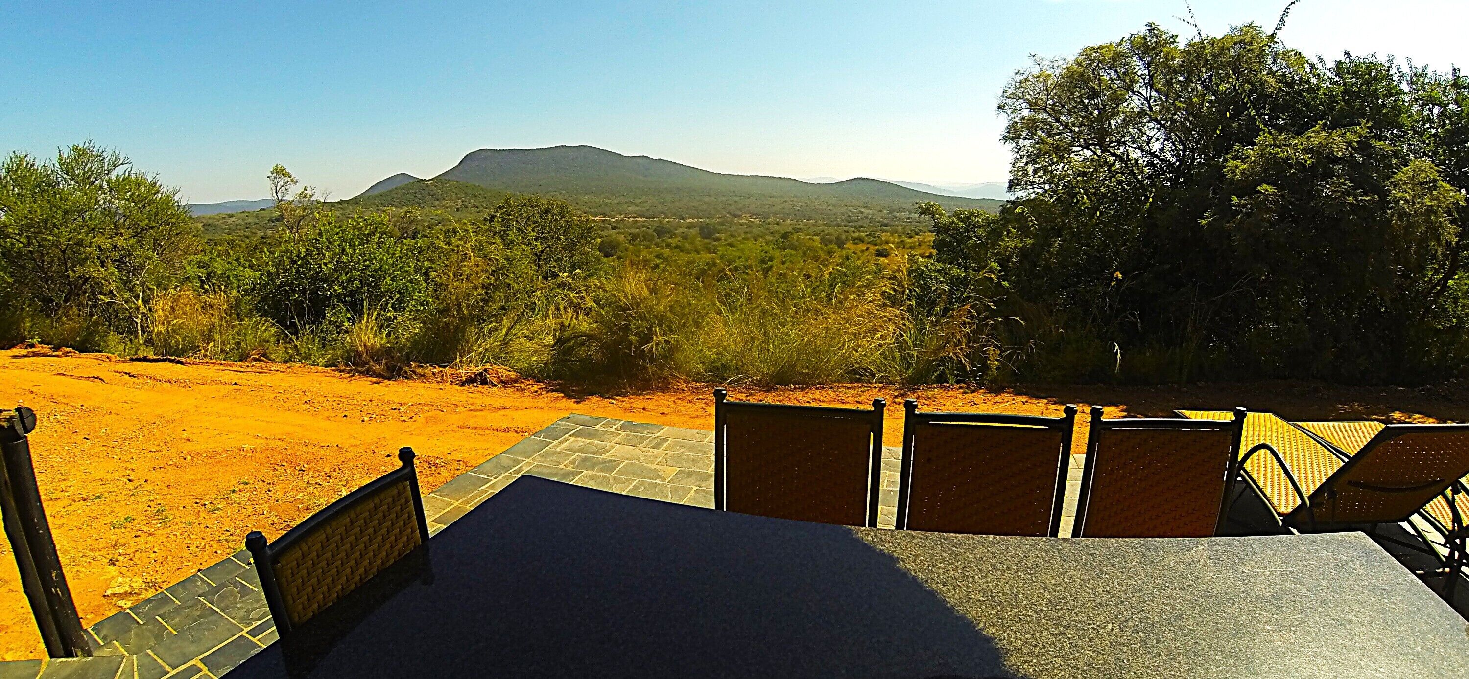 A terrace with a view of a mountain range, surrounded by greenery and a dirt path.