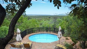 Zona de piscina con suelo de piedra, rodeada por una valla de metal negro y protegida por sombrillas.