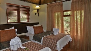 Habitación de hotel con dos camas, techo de paja, ventana con persianas, lámpara de pared y vista a zonas verdes.