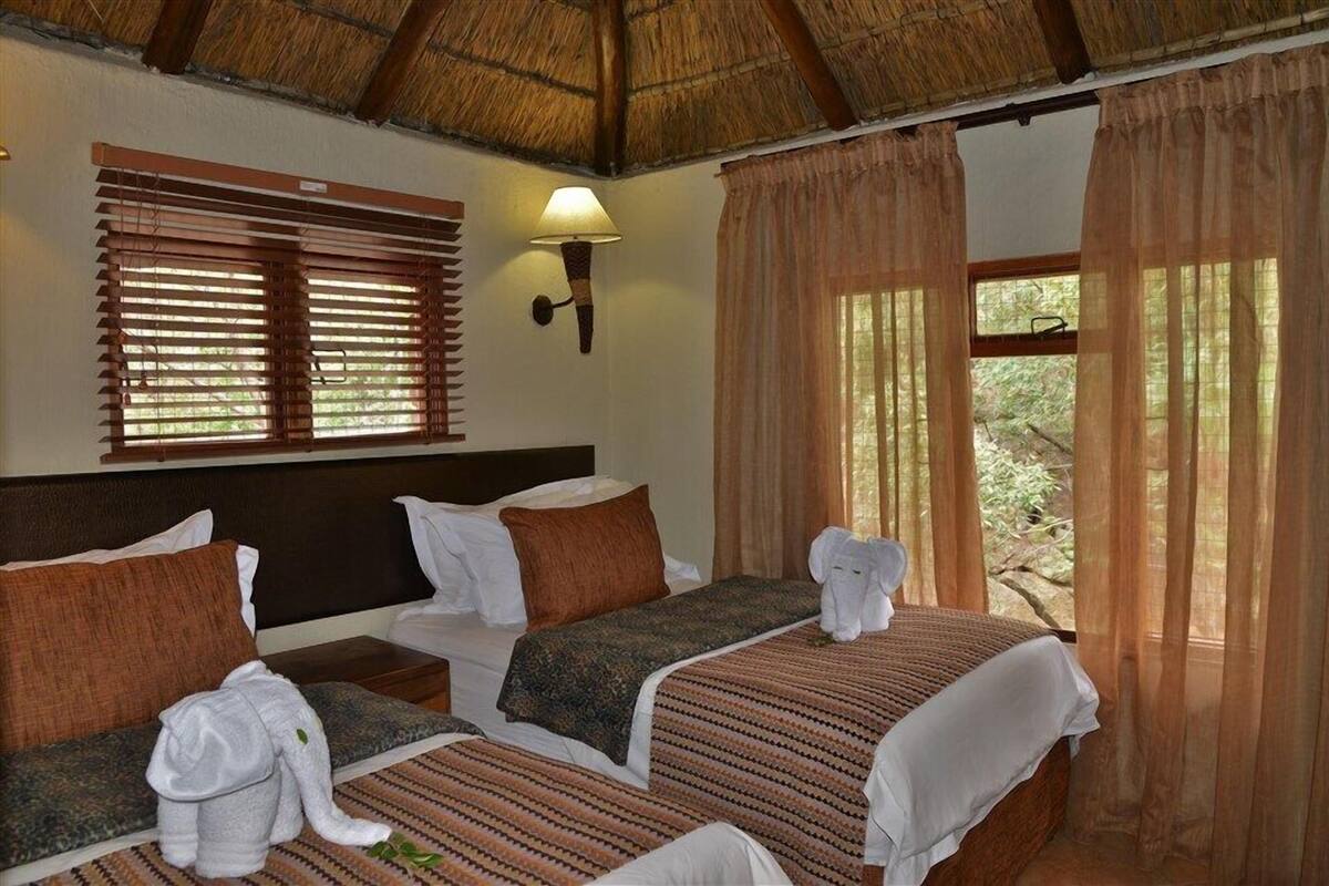 A hotel room with two beds, a thatched roof, a window with blinds, a wall-mounted lamp, and a view of greenery outside.