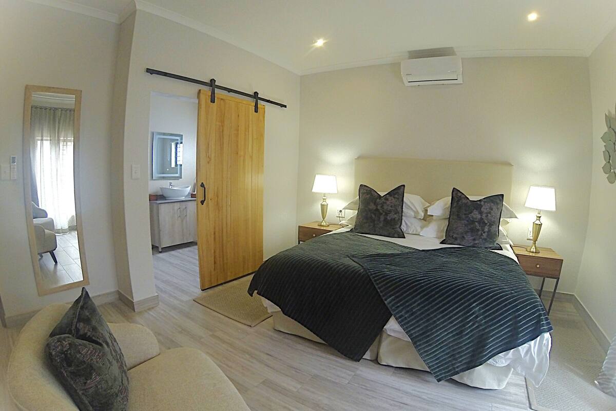 A bedroom with a bed, bedside tables, a nightstand, a chair, a bathroom with a sink and mirror, and a sliding door.