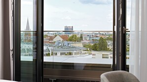 A hotel room with a large window overlooking a cityscape, including buildings and a church spire. - Numa Berlin Drift (Berlin)