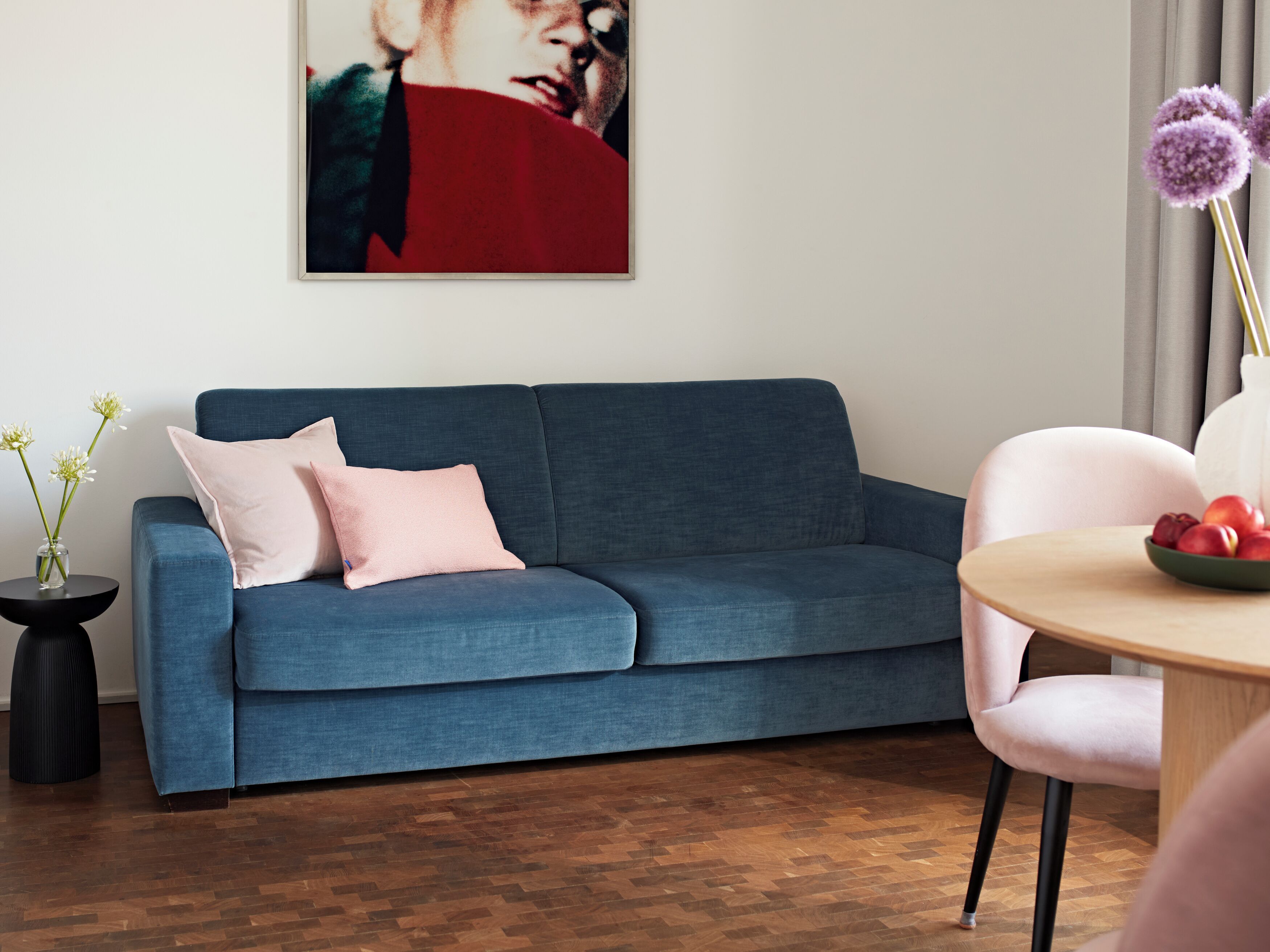 A modern living room with a blue sofa, a round wooden table with a bowl of fruit, and a vase with flowers.