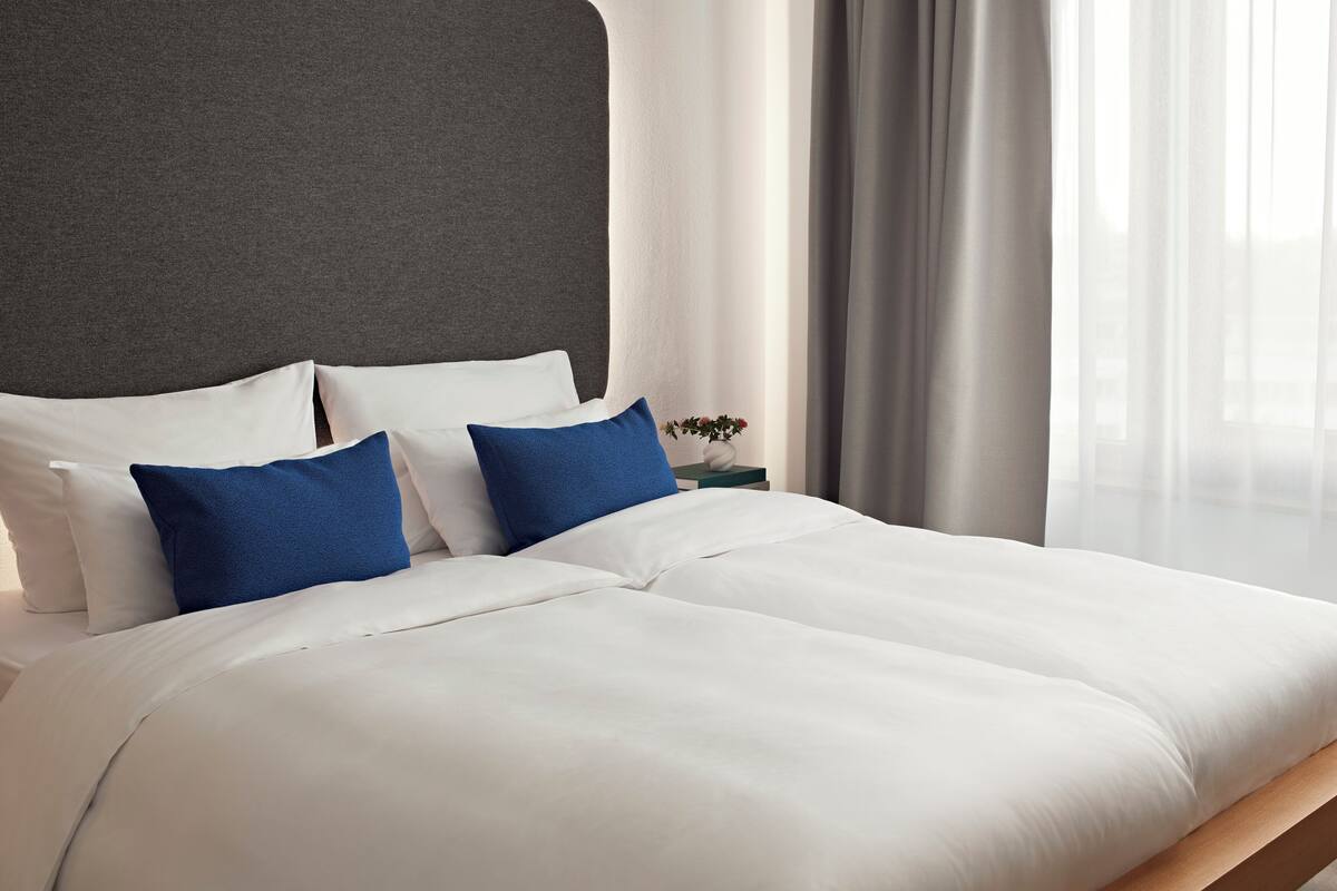 Una cama bien hecha con almohadas blancas y azules, un cabecero gris y una mesita de noche con un jarrón de flores.