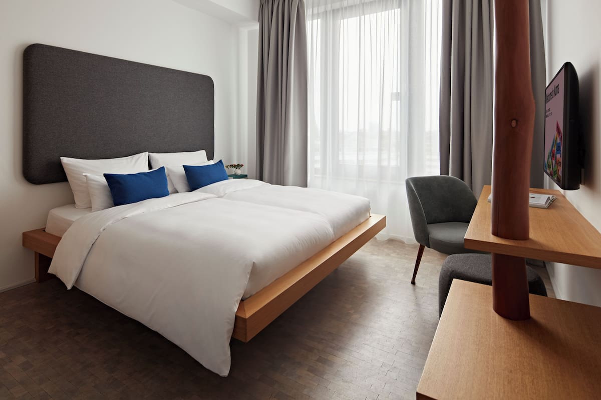 Una habitación de hotel moderna con una cama grande, una mesita de noche de madera, un escritorio y un televisor montado en la pared.