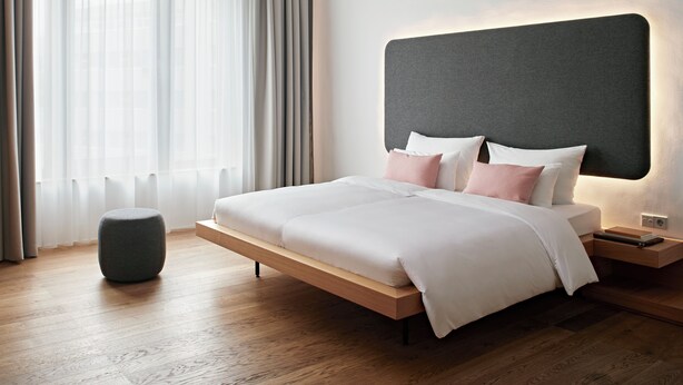 A modern bedroom with a large bed, a bedside table, a nightstand, a round ottoman, and a large window with sheer curtains. - Numa Berlin Drift (Berlin)