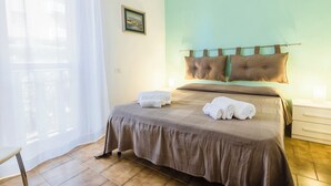 A neatly made bed with a brown headboard, a bedside table with a lamp, and a window with sheer curtains. - Da Milena (Orbetello)