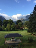 A gazebo with a table and chairs in a grassy area with trees and mountains in the background.