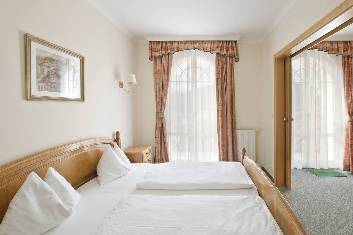 A hotel room with a double bed, a nightstand, a lamp, a framed picture on the wall, and a view of the outside through large windows with curtains.