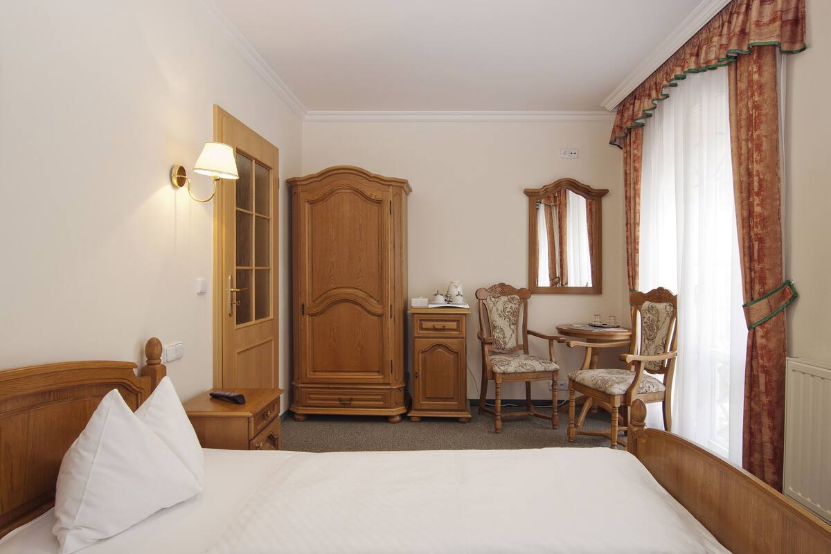 A hotel room with a bed, a wardrobe, a desk with a chair, a small table, and a mirror.