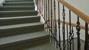 Una escalera con peldaños alfombrados de verde y una barandilla de madera, que conduce a una plataforma con una pared blanca y una lámpara.