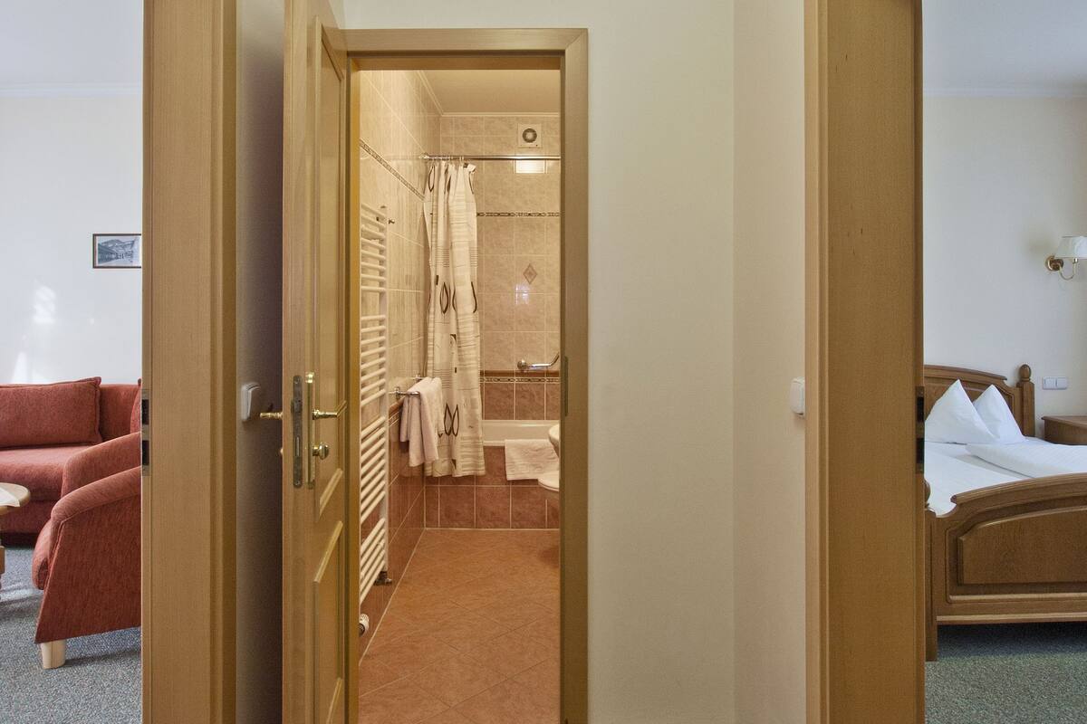 A hotel room with a bed, a bathroom with a shower, and a hallway leading to another room.