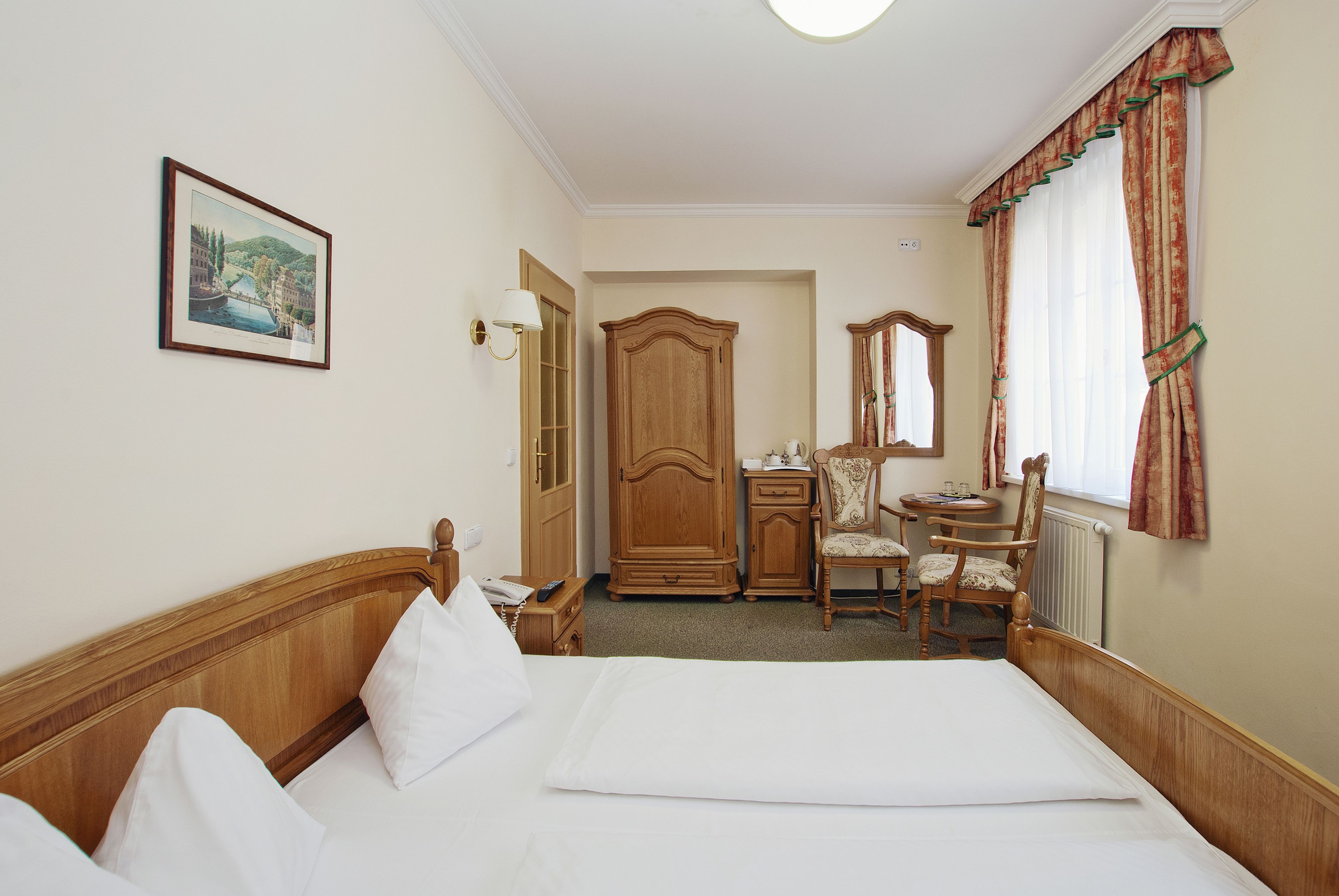 A hotel room with a large bed, a wooden wardrobe, a desk with a chair, a small table, and a window with curtains.