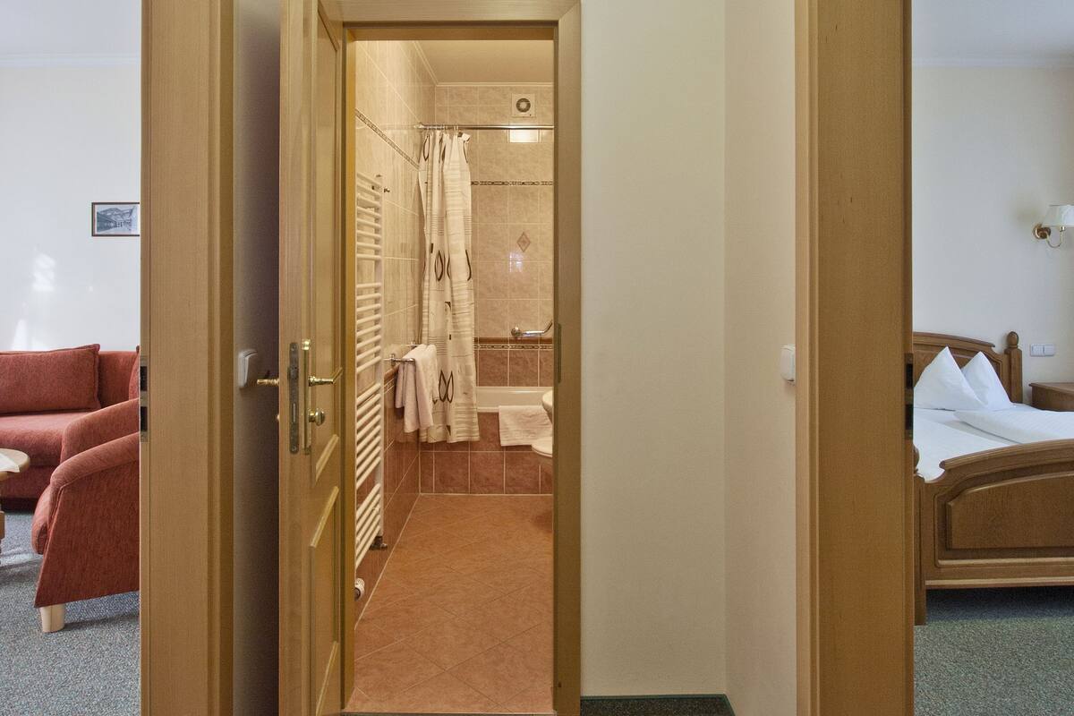 A hotel room with a bed, a bathroom with a shower, and a hallway leading to another room.