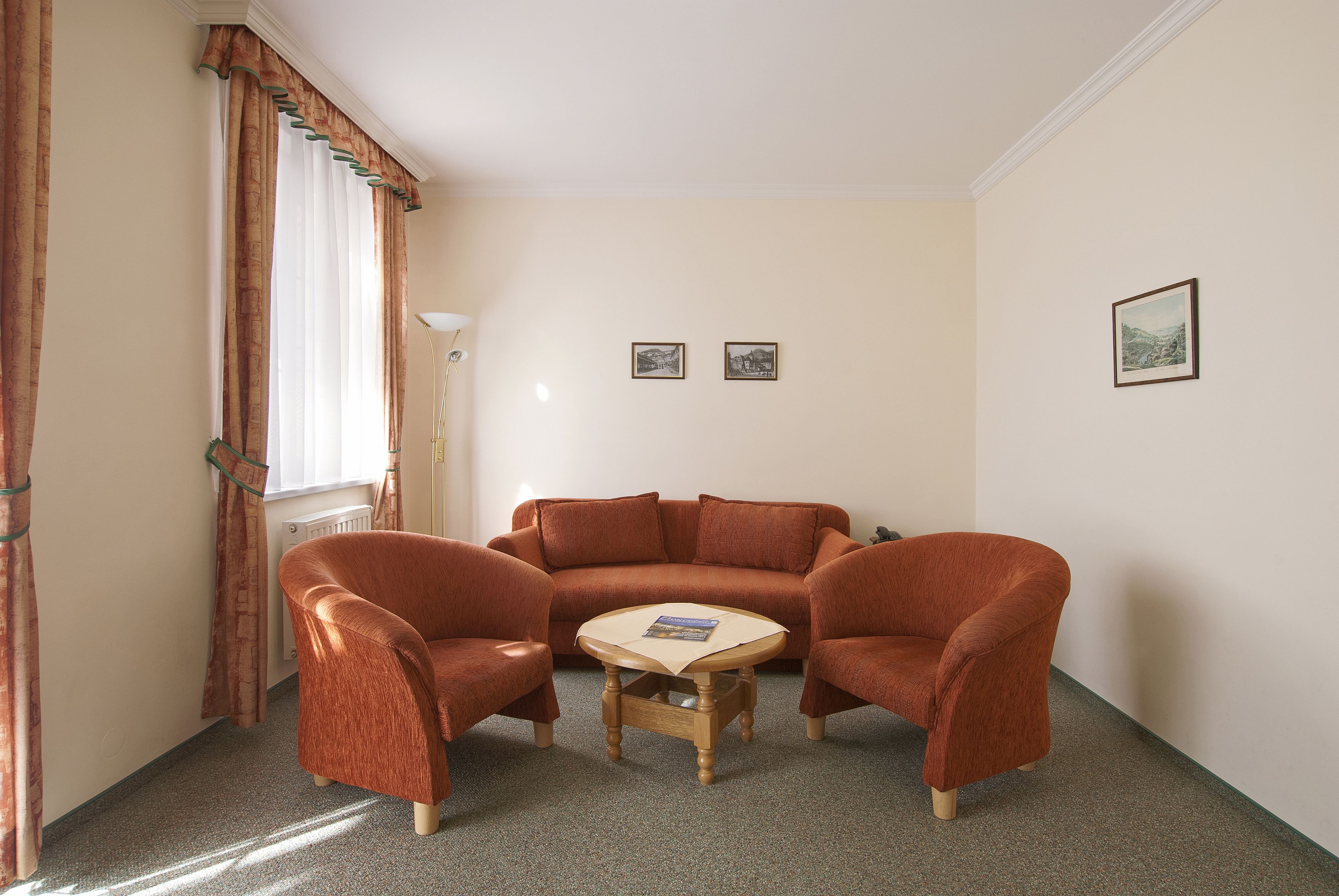 Una sala de estar con un sofá marrón, dos sillones y una pequeña mesa de centro de madera. Hay dos cuadros enmarcados en la pared y una ventana con cortinas.