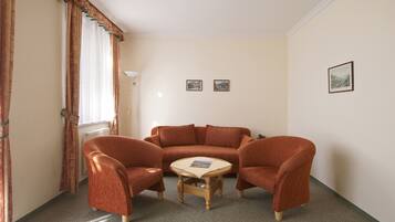 Una sala de estar con un sofá marrón, dos sillones y una pequeña mesa de centro de madera. Hay dos cuadros enmarcados en la pared y una ventana con cortinas.