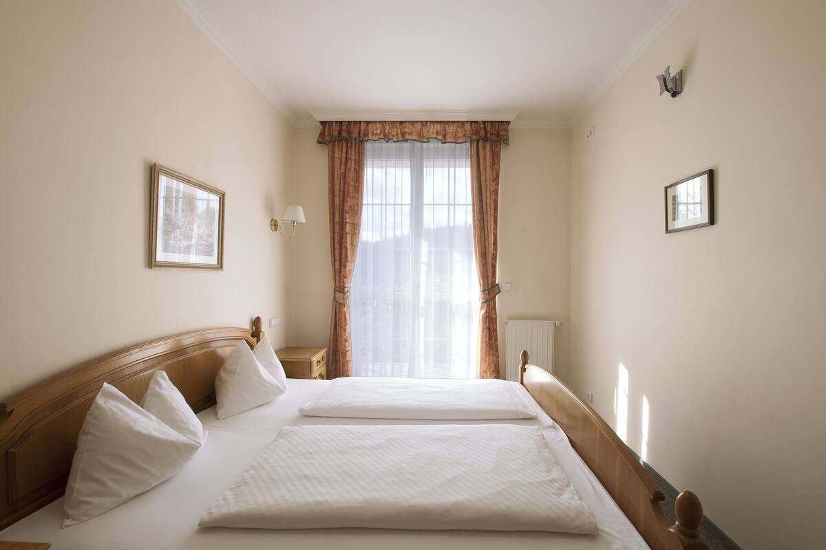 A hotel room with a large bed, wooden headboard, two bedside tables, a nightstand, and a window with curtains.