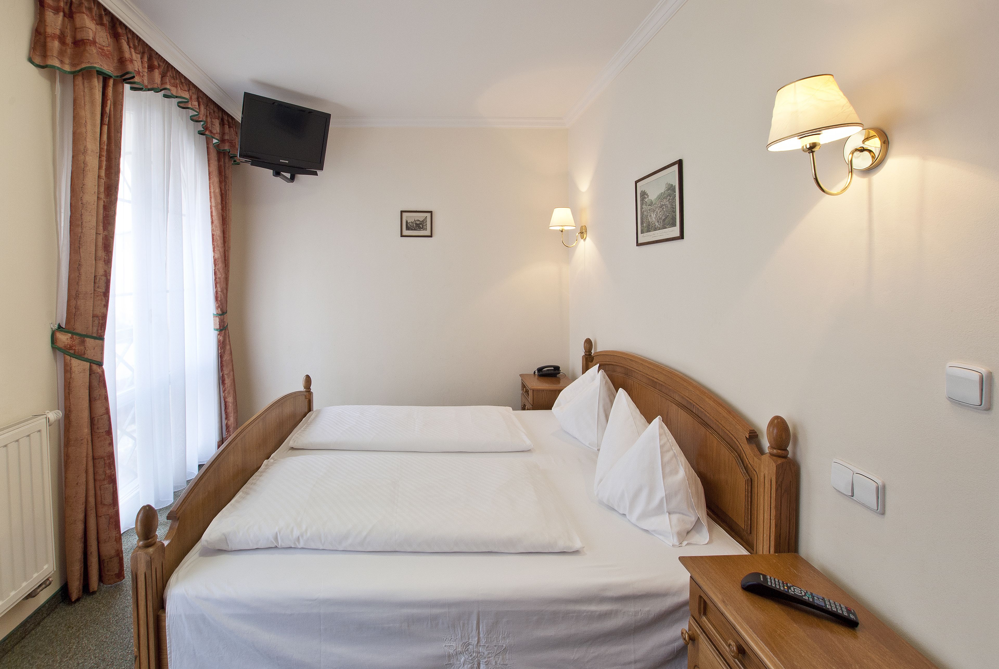 A hotel room with a double bed, a bedside table with a remote control, a wall-mounted TV, and two wall sconces.