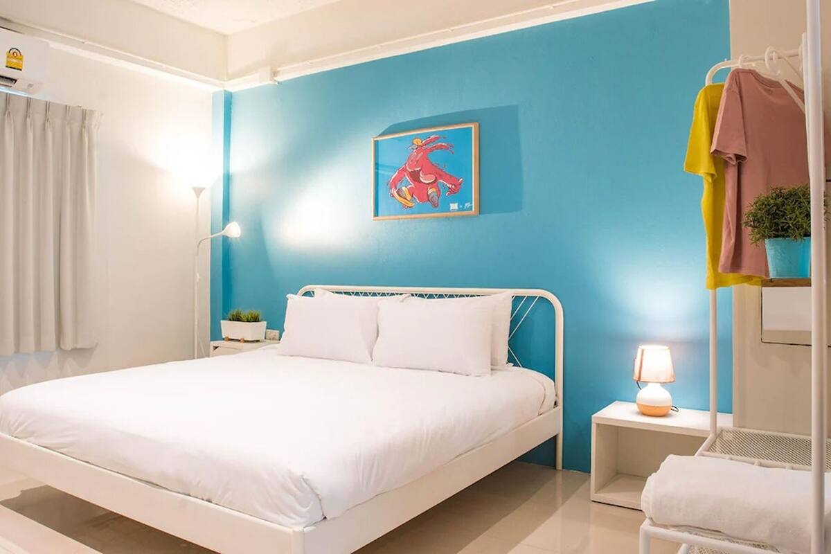 Ein Schlafzimmer mit einem weißen Bett, einer blauen Wand, einem gerahmten Bild, einer Nachttischlampe und einer kleinen Pflanze.
