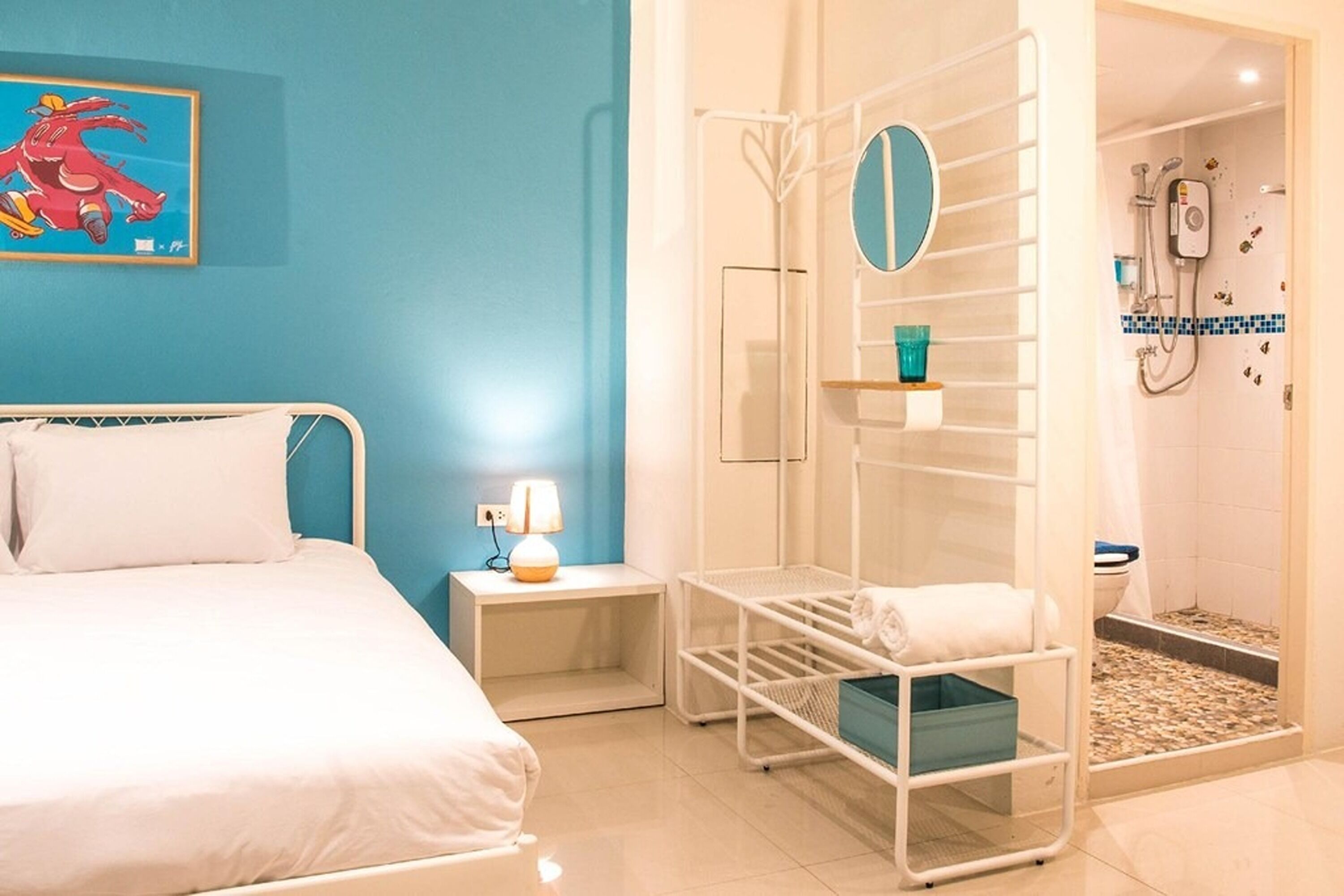 A hotel room with a bed, a nightstand with a lamp, a drying rack, and a shower area.
