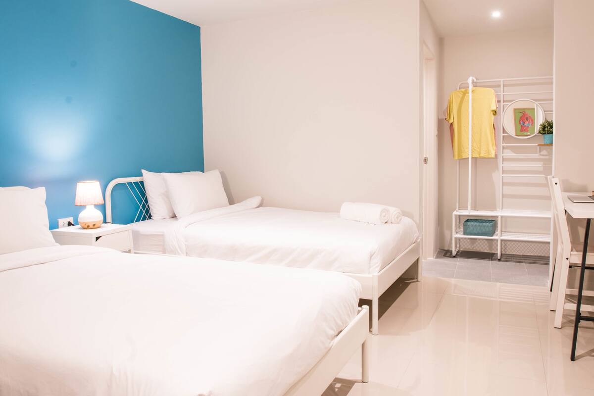 Ein Hotelzimmer mit zwei Betten, einem Schreibtisch, einem Stuhl, einer Lampe, einem Regal mit einer Pflanze und einer Treppe.