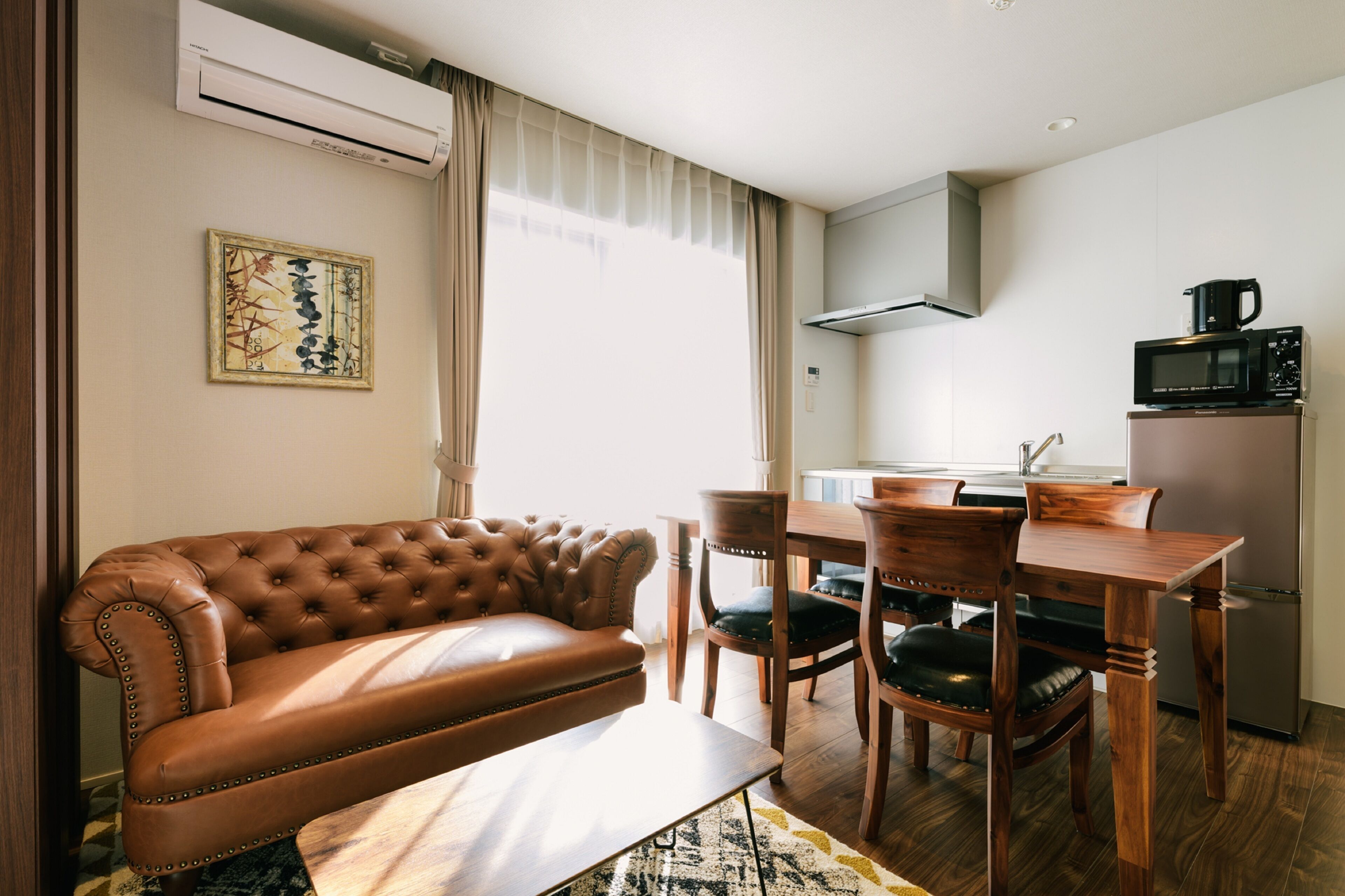 A modern living room with a leather sofa, wooden dining table, and chairs. There is a kitchen area with a microwave and a refrigerator.