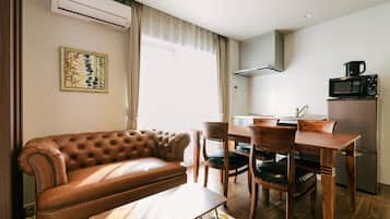 A modern living room with a leather sofa, wooden dining table, and chairs. There is a kitchen area with a microwave and a refrigerator.