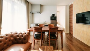 Una sala moderna con un sofá de cuero, una mesa de comedor de madera y sillas. Se ve un área de cocina con microondas y estufa al fondo.