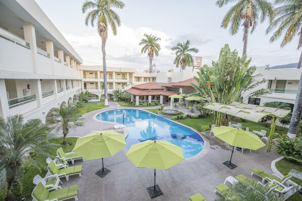 Un área de piscina con sillas de descanso y sombrillas, rodeada de palmeras y un edificio de varios pisos.